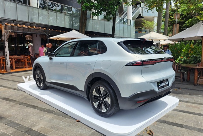 Silver electric SUV on a white display platform at an outdoor showroom, with people and umbrellas in the background.